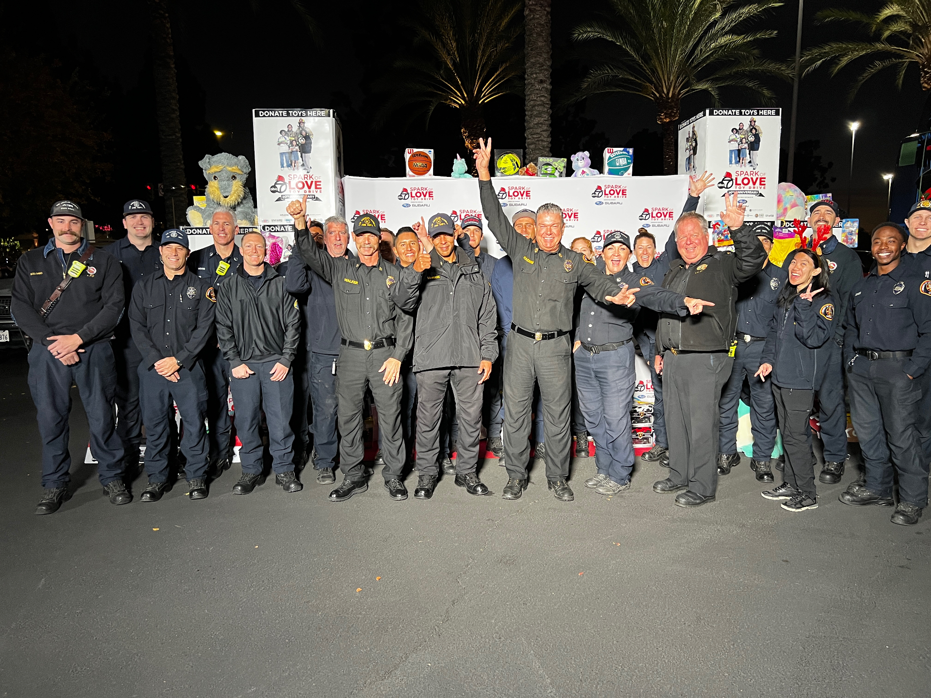 Firefighters posing in front of Spark of Love Toy Drive donation boxes at nighttime ABC7 event